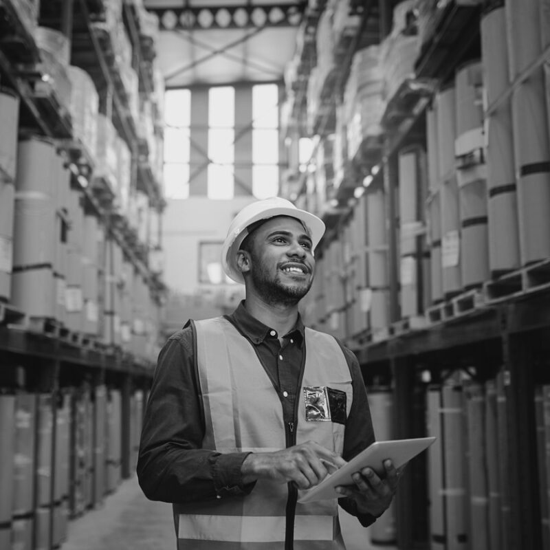 Portrait of young man wearing safety jacket holding digital tablet standing in factory warehouse