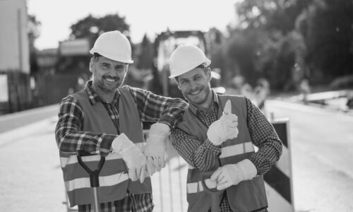 Two construction workers in hardhats and vests posing for a photo at the road