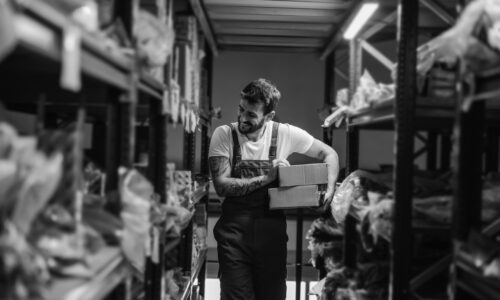 Hardworking smiling tattooed bearded worker relocating boxes while walking in storage of import and export firm.