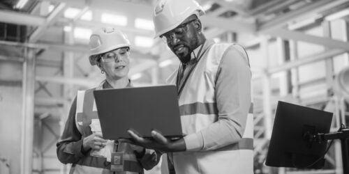 Team of Diverse Professional Heavy Industry Engineers Wearing Safety Uniform and Hard Hats Working on Laptop Computer. African American Technician and Female Worker Talking on a Meeting in a Factory.