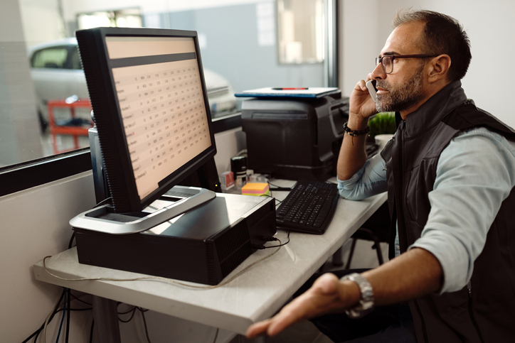 Foreman of auto mechanic workshop talking on the phone while using computer in the office