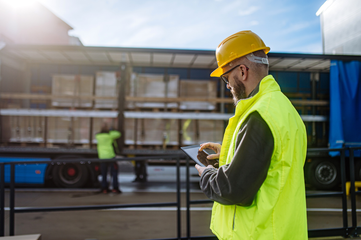 Warehouse manager overseeing unloading of truck, holding tablet, looking at cargo details, checking delivered items, goods agaist order