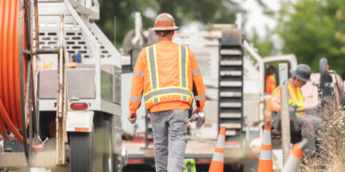 A construction worker walks along the road to heavy equipment that's parked alongside the area they're working on. He is carrying a can of spray paint and measuring tape as he walks away from the camera.