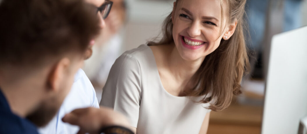 Smiling happy young woman talking with male colleagues at shared workplace, worker laughing at funny joke, reading funny news, excited business success, employees having break, pause