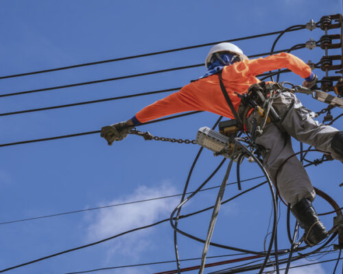 Low angle view of electrician with safety equipment and various work tools is installing cable lines and electrical system on electric power pole against blue sky background