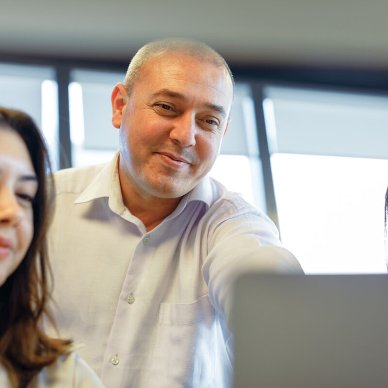Joyful Collaboration: Manager Guides Young Professionals in Modern Office. Immerse yourself in the vibrant energy of a modern office as a team of happy business people gathers around a computer, their faces beaming with enthusiasm. The middle-aged manager, a seasoned boss, takes center stage, passionately showing and explaining valuable insights on the screen to two young, beautiful businesswomen or interns. Witness the exchange of information and knowledge flow seamlessly among working business employees, creating an atmosphere of shared success and growth in the workplace