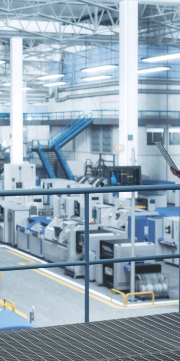 Senior Engineer Standing on a Platform, Using Laptop Computer at an Electronics Factory. Machines are Undergoing Maintenance, Specialist Monitoring the Progress Through Online Software
