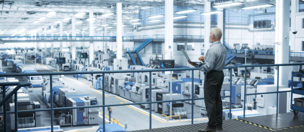 Senior Engineer Standing on a Platform, Using Laptop Computer at an Electronics Factory. Machines are Undergoing Maintenance, Specialist Monitoring the Progress Through Online Software