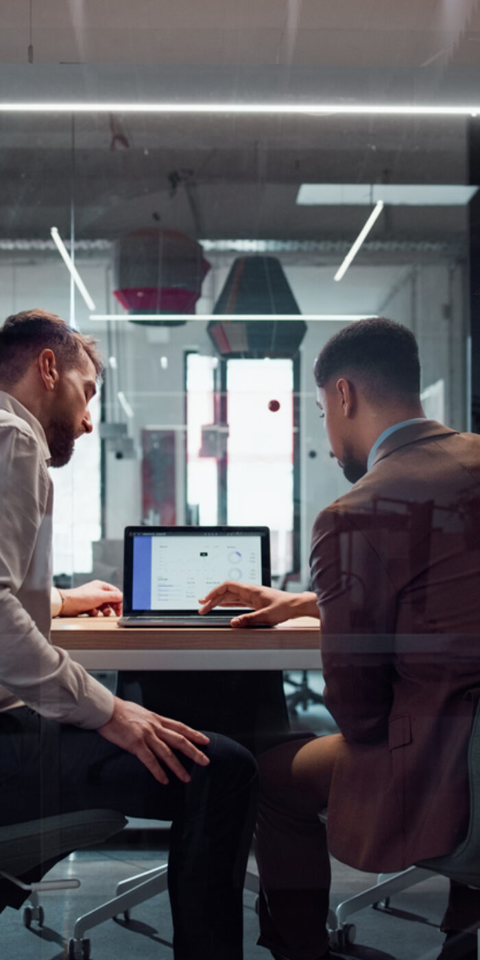 A group of coworkers discuss ideas at a laptop in a modern glass-walled workspace, emphasizing teamwork and professional collaboration. The office fosters creativity and productivity with its open and interactive environment.