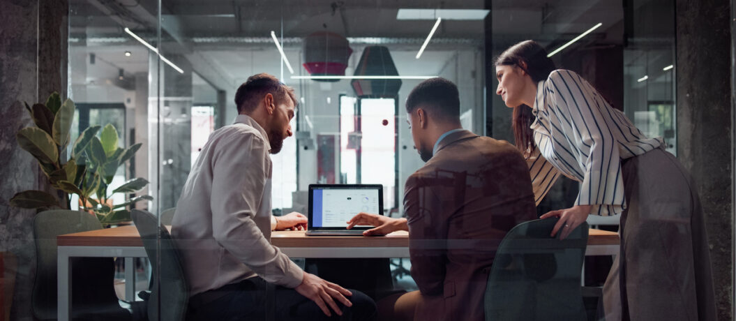 A group of coworkers discuss ideas at a laptop in a modern glass-walled workspace, emphasizing teamwork and professional collaboration. The office fosters creativity and productivity with its open and interactive environment.