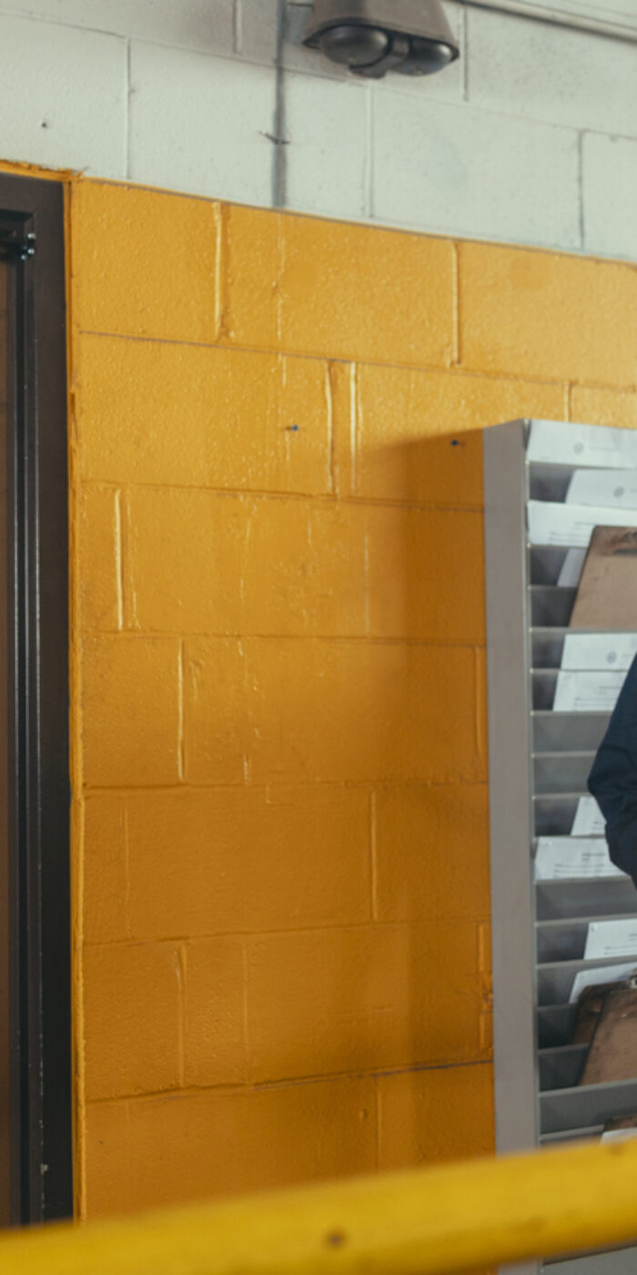Man wearing blue coveralls holding a clipboard and looking down, standing next to a yellow brick wall and an employees only door in an industrial garage