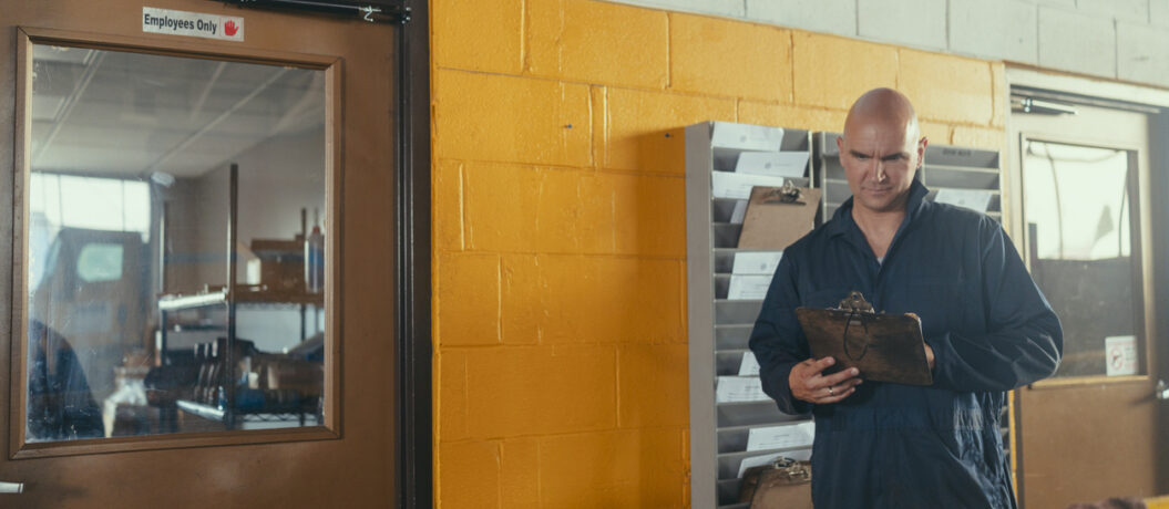 Man wearing blue coveralls holding a clipboard and looking down, standing next to a yellow brick wall and an employees only door in an industrial garage