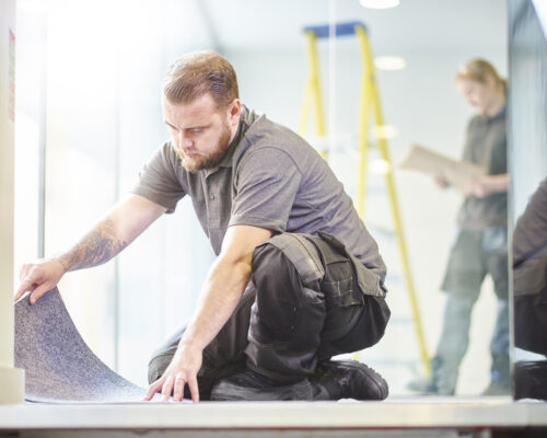 a male carpet fitter is installing carpet  in a modern office refurbishment. IN the background a female co-worker can be seen holding plans . They are wearing safety workwear.