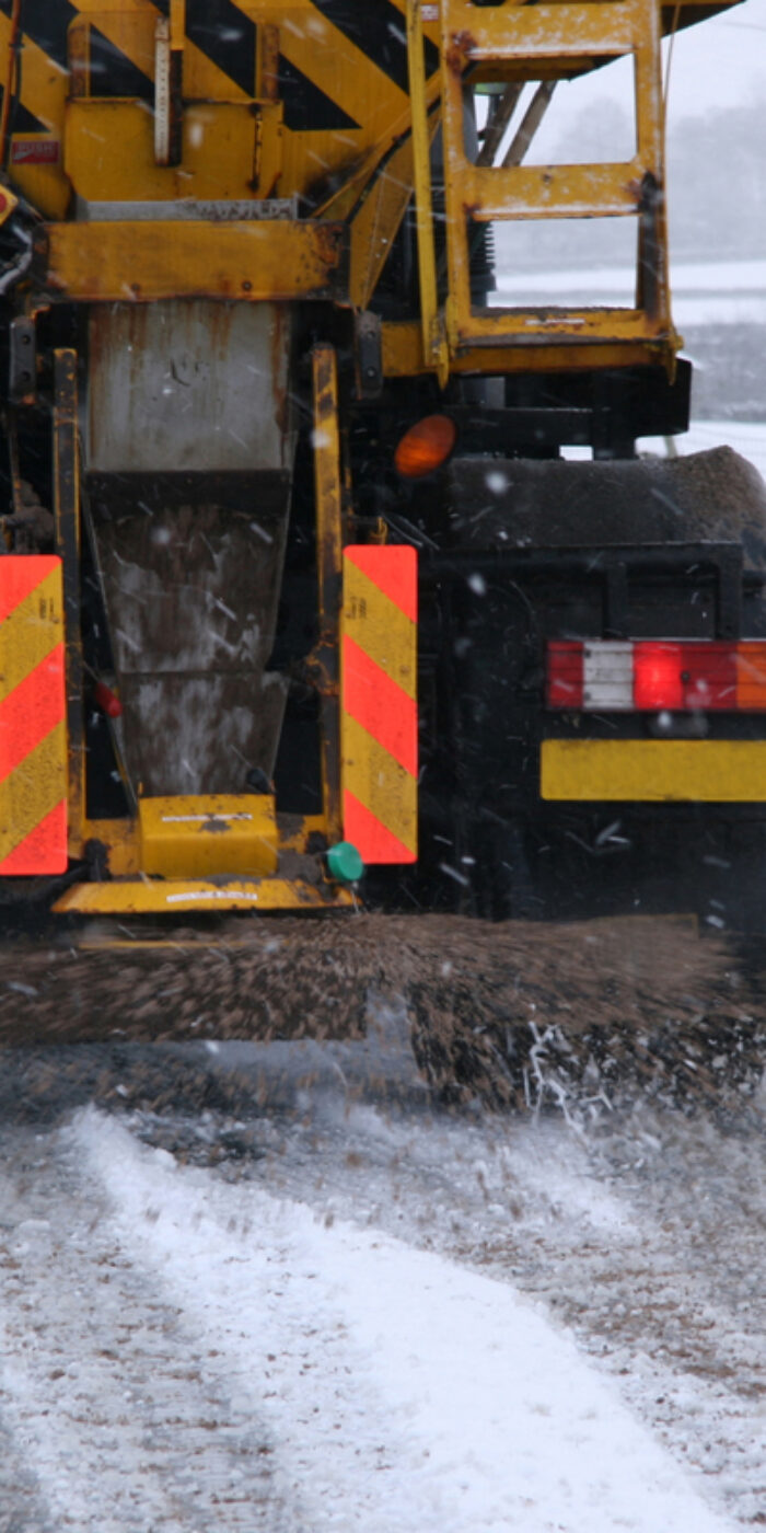 Snow plough gritting the roads in the Yorkshire Dales, UK. Road is covered in snow. Gritting truck is moving down an empty road whilst gritting. Stone wall and fence can be seen.