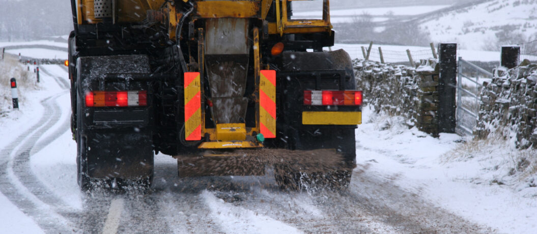 Snow plough gritting the roads in the Yorkshire Dales, UK. Road is covered in snow. Gritting truck is moving down an empty road whilst gritting. Stone wall and fence can be seen.