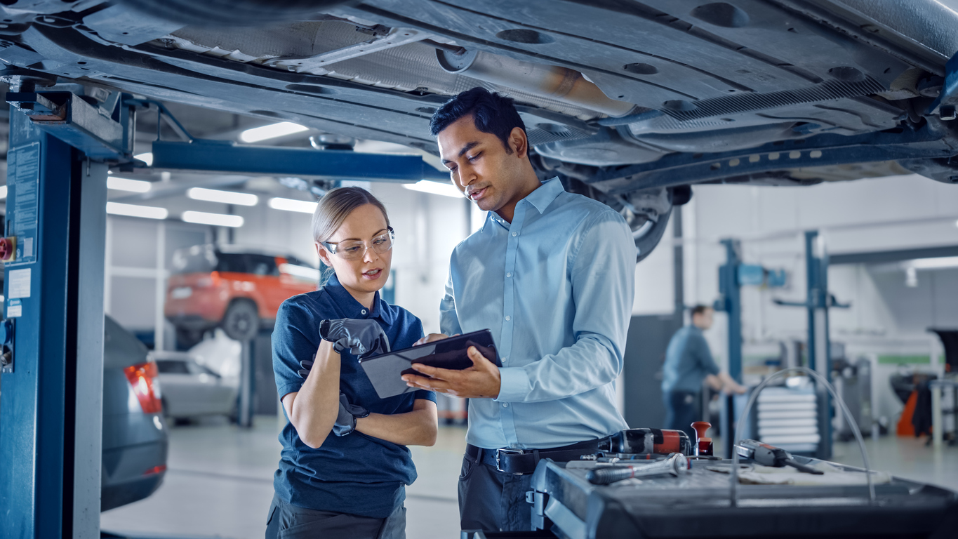 Female Mechanic Talking to a Manager Under a Vehicle in a Car Service. Specialist is Showing Info on a Tablet Computer. Empowering Woman Wearing Gloves and Safety Gloves. Modern Clean Workshop. preventive maintenance