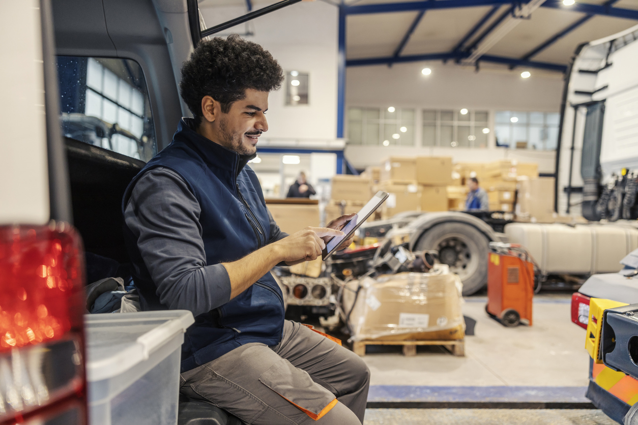 Interracial trucking company worker sitting on vehicle and scrolling on tablet
