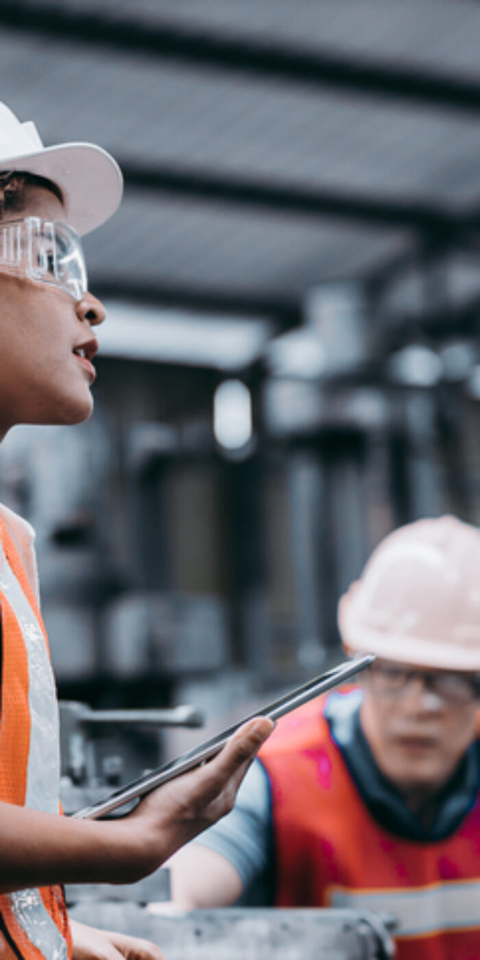 Female industrial engineer wearing a white helmet while standing in a heavy industrial factory behind she talking with workers, Various metal parts of the project