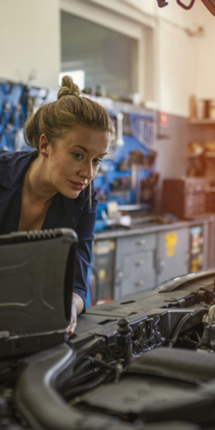 An attractive woman mechanic working on a car in a repair shop. A female mechanic is working under a bonnet of a car in a garage repair shop. She is wearing blue overalls.