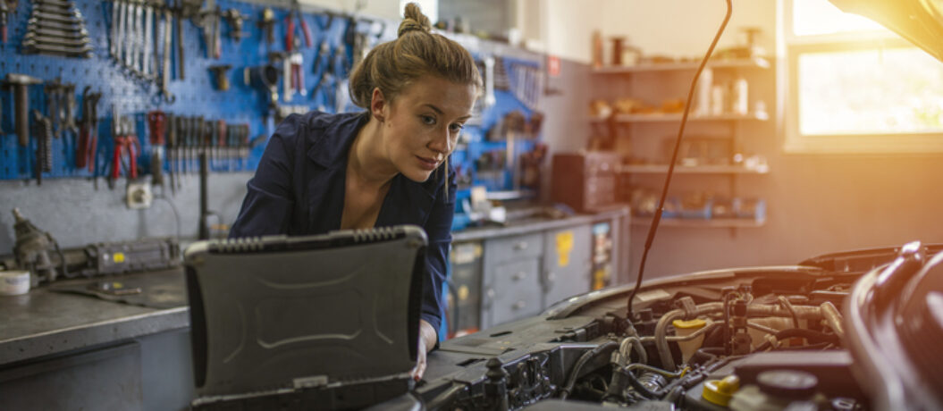 An attractive woman mechanic working on a car in a repair shop. A female mechanic is working under a bonnet of a car in a garage repair shop. She is wearing blue overalls.