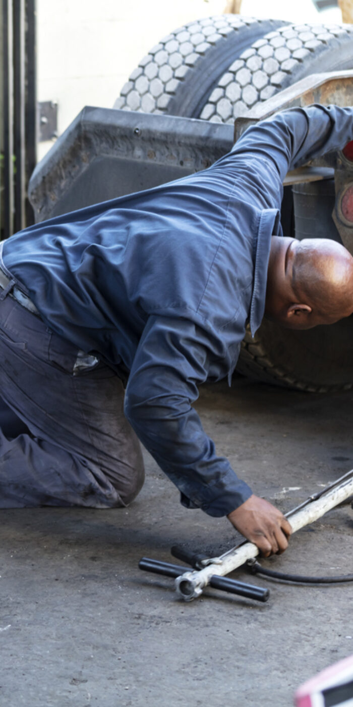 A mature African-American man in his 40s working in a truck repair shop. He is kneeling on the floor, looking under the rear of the truck, sliding a jack underneath.