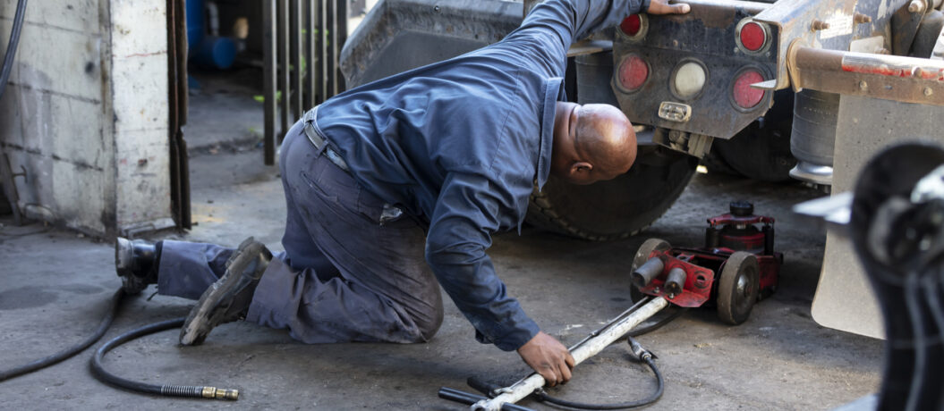 A mature African-American man in his 40s working in a truck repair shop. He is kneeling on the floor, looking under the rear of the truck, sliding a jack underneath.