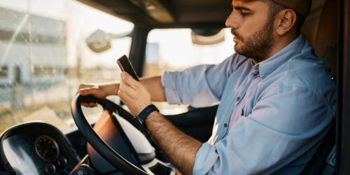 Young truck driver using mobile phone while driving transport vehicle.