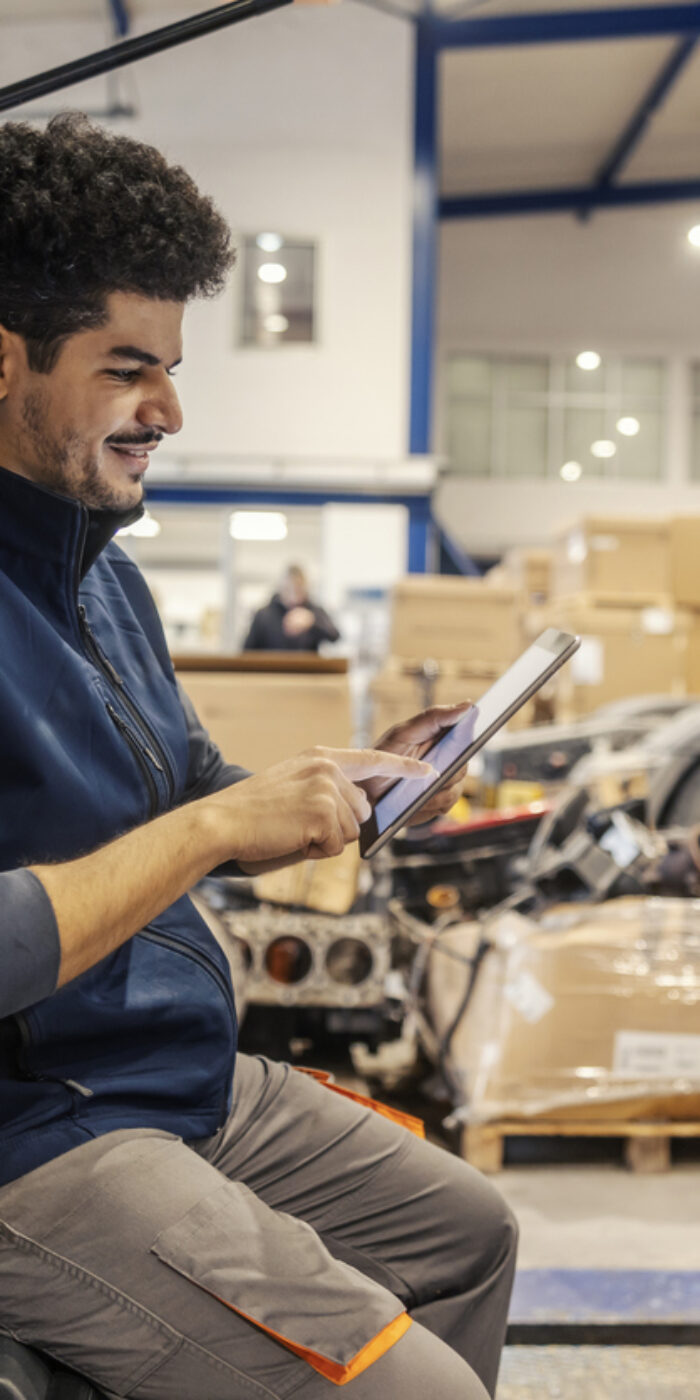 Side view of multicultural trucking company workman sitting on vehicle in facility and scrolling on tablet.