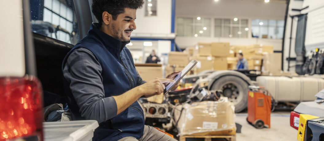 Side view of multicultural trucking company workman sitting on vehicle in facility and scrolling on tablet.