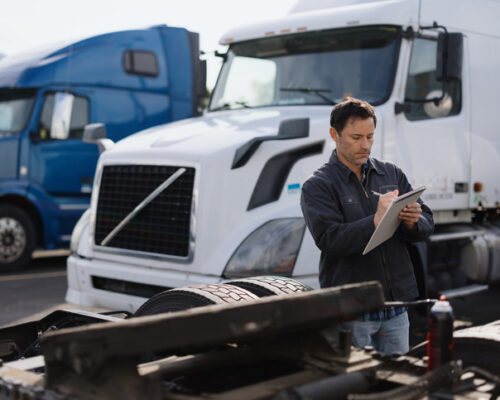 Truck driver writing notes while inspecting a semi-truck trailer in a parking lot, with other trucks in the background