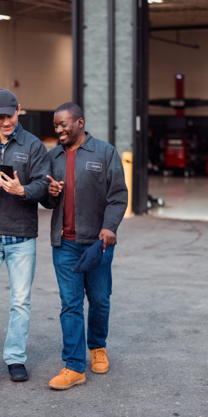 Two male mechanics enjoy a cheerful conversation outside a repair garage. Dressed in work jackets, they share a friendly mood in a professional setting, reflecting teamwork and camaraderie.