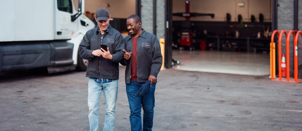 Two male mechanics enjoy a cheerful conversation outside a repair garage. Dressed in work jackets, they share a friendly mood in a professional setting, reflecting teamwork and camaraderie.