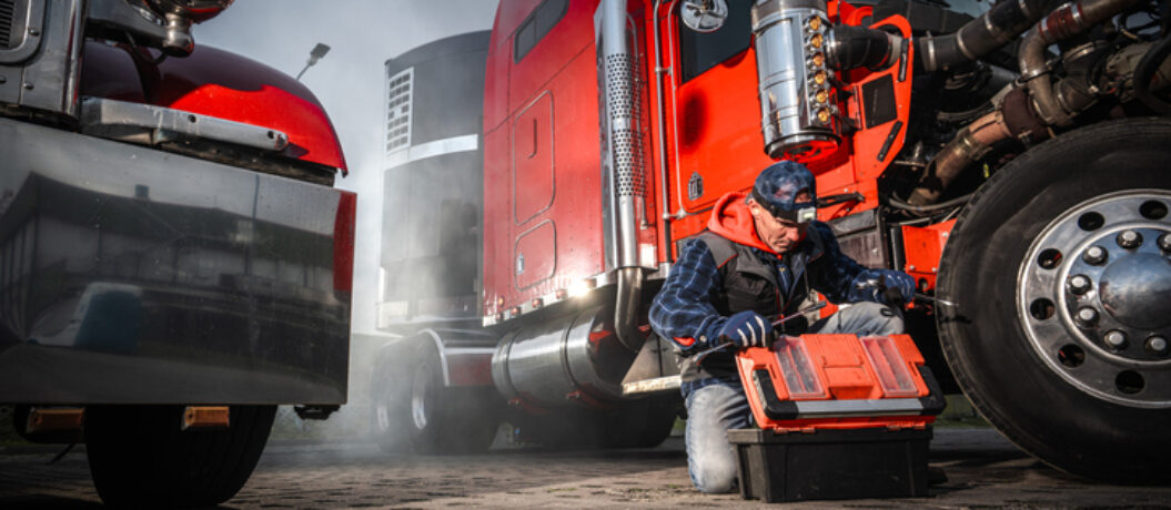 A mechanic crouches next to a large red truck, examining tools in a black toolbox. Smoke rises from the engine in an urban environment.