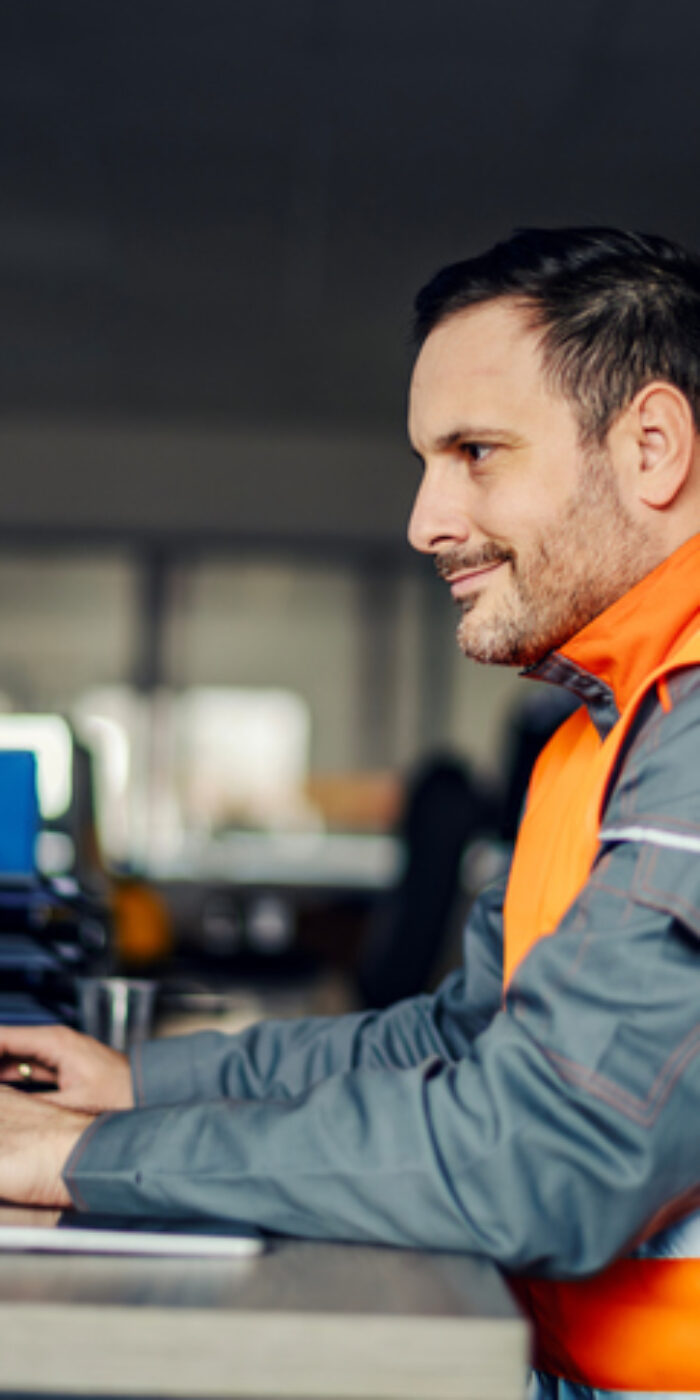 Industrial worker wearing safety vest and uniform, sitting at a desk and typing on a computer keyboard, managing logistics and operations in an office environment
