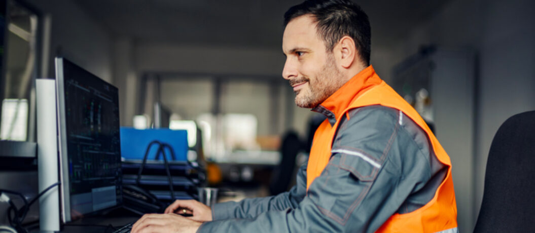 Industrial worker wearing safety vest and uniform, sitting at a desk and typing on a computer keyboard, managing logistics and operations in an office environment
