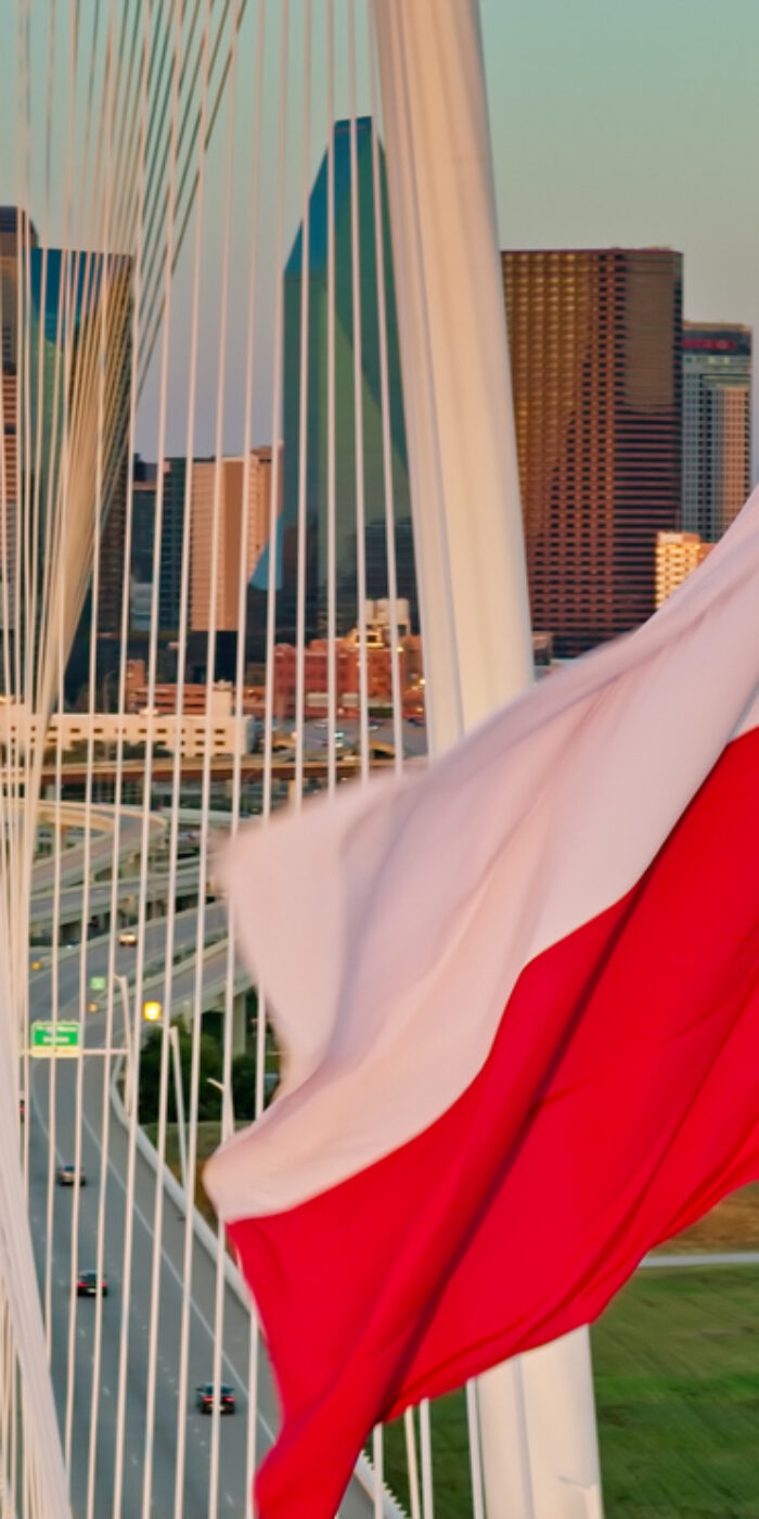 A Texas state flag waves prominently in the foreground as the Margaret Hunt Hill Bridge spans the Trinity River with downtown Dallas rising in the background during golden hour. The cable-stayed bridge, designed by Santiago Calatrava, connects West Dallas to the central business district. The downtown skyline features numerous high-rise buildings and skyscrapers illuminated by warm afternoon light. This aerial perspective captures the iconic white arch and cable system of the bridge with vehicular traffic flowing across its deck, while the Trinity River and surrounding parkland are visible below.   • Authorization was obtained from the FAA for this operation in restricted airspace.
