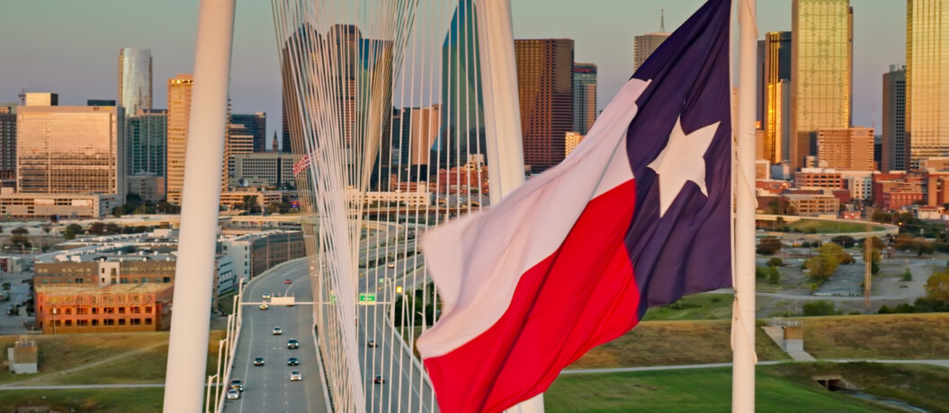 A Texas state flag waves prominently in the foreground as the Margaret Hunt Hill Bridge spans the Trinity River with downtown Dallas rising in the background during golden hour. The cable-stayed bridge, designed by Santiago Calatrava, connects West Dallas to the central business district. The downtown skyline features numerous high-rise buildings and skyscrapers illuminated by warm afternoon light. This aerial perspective captures the iconic white arch and cable system of the bridge with vehicular traffic flowing across its deck, while the Trinity River and surrounding parkland are visible below.   • Authorization was obtained from the FAA for this operation in restricted airspace.