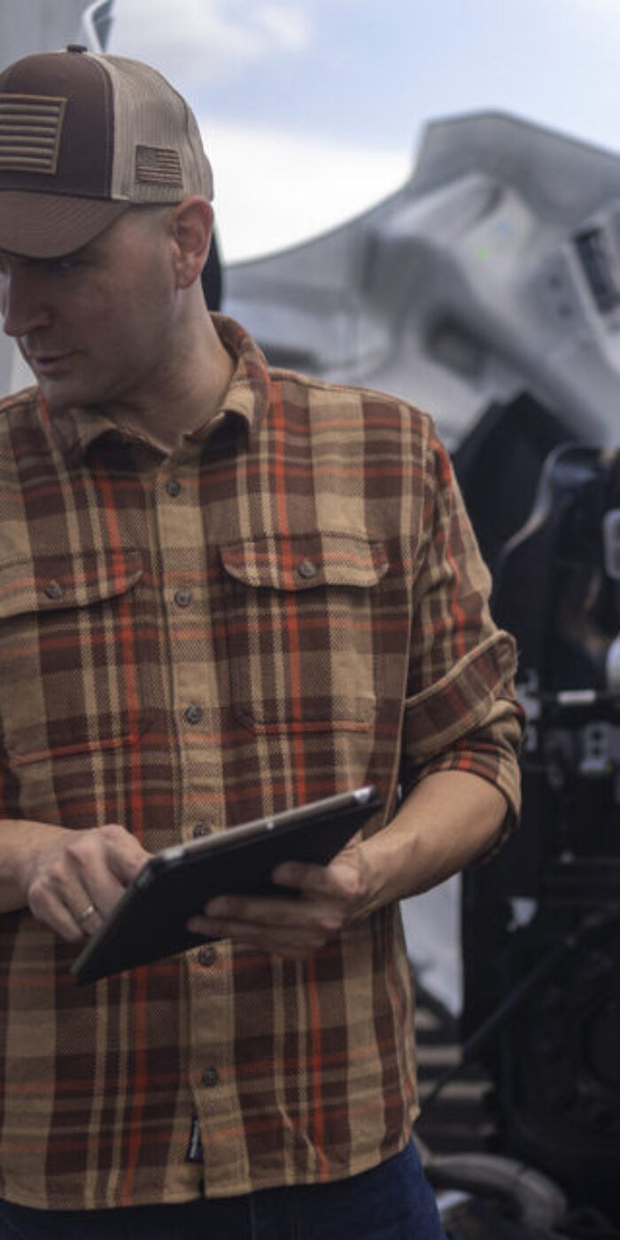 A bearded auto mechanic in a grey hoodie and a truck driver in a plaid shirt standing by the open hood of a semi-truck. The driver is holding a digital tablet, likely reviewing diagnostic data