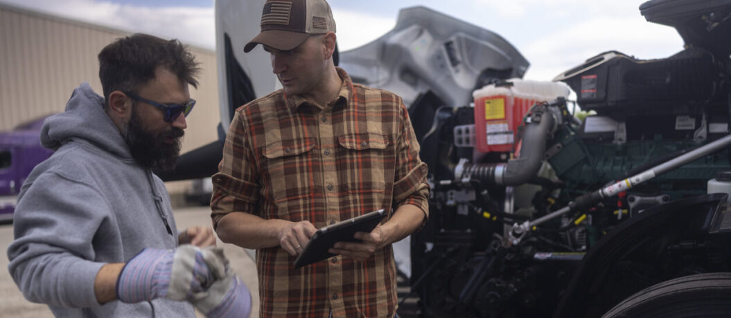 A bearded auto mechanic in a grey hoodie and a truck driver in a plaid shirt standing by the open hood of a semi-truck. The driver is holding a digital tablet, likely reviewing diagnostic data